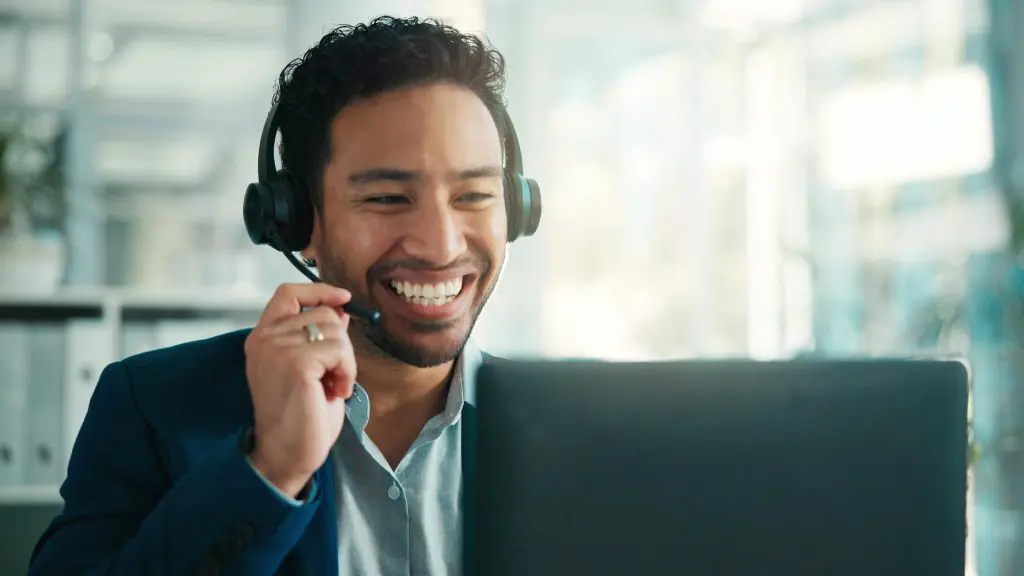 Smiling customer support agent using a headset and laptop to manage calls with modern VoIP phone systems in a professional office setting.